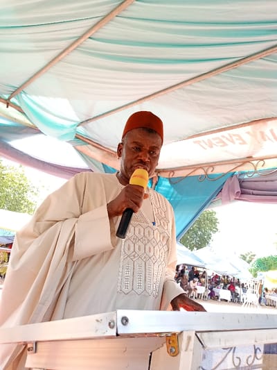 A middle-aged man wearing a white robe and a red hat is speaking into a yellow microphone. He is standing behind a wooden podium with a microphone stand. He is speaking to a crowd of people in the background. The scene is outdoors, possibly at a public event or ceremony. The location is Zum, Nigeria, at the coordinates 10.116 latitude and 13.046 longitude. The background features a blue and white canopy tent, trees, and other attendees at the event.