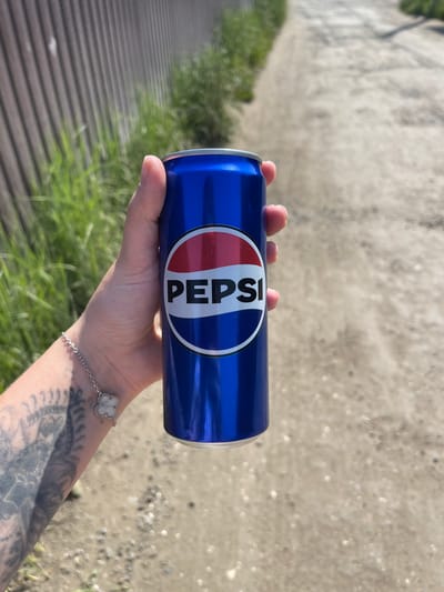 A person's hand holds a blue Pepsi can against a blurred background of a dirt path and a wooden fence. The Pepsi logo is clearly visible on the can. The person is wearing a bracelet with a clover-shaped charm. Tattoos are visible on the person's arm. The scene appears to be outdoors, possibly in a rural or suburban setting. No specific location in Molokovo, Russia, can be determined from the image.
