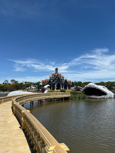 The image shows a view of the Ancient City (Muang Boran) in Bang Pu Subdistrict Municipality, Thailand. A large, open-mouthed crocodile sculpture is partially submerged in the water. A decorative Thai-style pavilion with layered roofs is visible in the background. A golden-colored bridge with detailed railings leads toward the pavilion. The sky is blue with scattered cirrus clouds. The water appears still. Vegetation lines the water's edge.