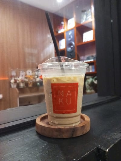 A clear plastic cup, filled with a light-colored iced beverage and containing a black straw, stands centered on a round wooden coaster. The cup exhibits condensation droplets on its exterior and features a red square label with "NAKU" inscribed in white text. The coaster and cup rest on a dark, possibly black, flat surface, which also shows moisture. The background, viewed through a reflective barrier, reveals an indoor setting. To the left, a light-brown wall or paneling is visible, with blurred reflections of small, decorative items on shelves. To the right, a darker wooden shelving unit holds various objects, notably a prominent dark audio speaker. The environment is illuminated by interior lighting. This scene is located at NAKU Kopi & Co. in Pontianak, Indonesia. No individuals, direct actions, or interactions are visible.