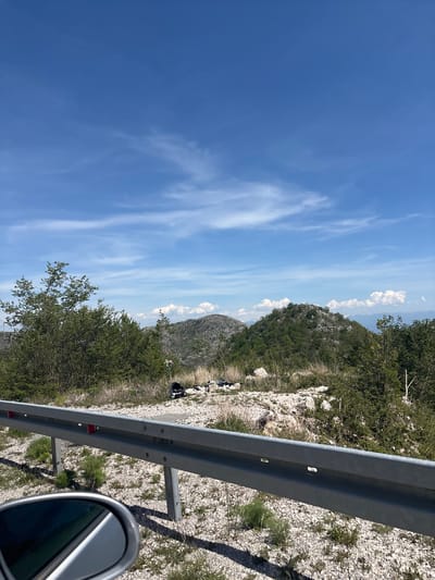 This image captures a bright, sunny mountainous landscape, likely viewed from a vehicle on a road. The scene is dominated by natural elements, with clear blue skies and rugged terrain.

In the immediate foreground, a metallic silver guardrail runs horizontally across the bottom right quadrant of the image, indicating the edge of a roadway or a scenic overlook. A portion of a car's side mirror is visible in the bottom right corner, reflecting the guardrail and the sky, suggesting the perspective is from inside a car.

Beyond the guardrail, the ground is covered in light-colored, rocky scree and sparse green vegetation, consistent with scrubland or mountainous slopes. Further into the mid-ground, a larger hill or mountain rises, densely covered in green trees and bushes. In the distance, several layers of hills and mountains recede, partially obscured by the clear air. Near the base of the mid-ground hill, behind the guardrail, some dark, unidentifiable objects or forms are visible on the ground.

The sky is a vibrant blue, stretching across most of the upper and left portions of the image, adorned with scattered white, wispy clouds. The lighting suggests it is bright daylight. The overall scene conveys a sense of open space and natural beauty, characteristic of mountainous regions like Obzovica, Montenegro. No visible text is present in the image.
