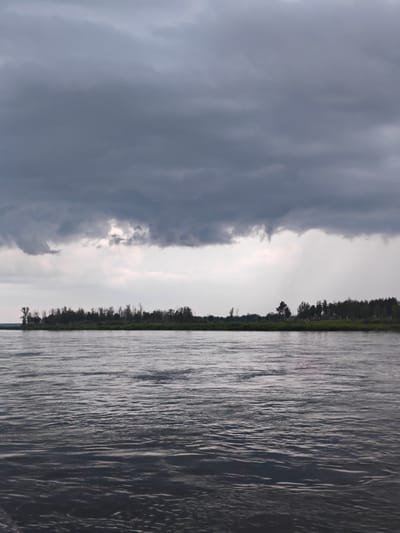The image depicts a wide, calm river flowing through a natural landscape. The sky above is heavily overcast with dark, dramatic storm clouds. A treeline with a mix of deciduous and coniferous trees borders the far bank of the river. The water's surface reflects the muted tones of the sky and surrounding vegetation. A faint indication of rain can be observed in the distance, falling over the treeline. The overall scene suggests a pre-storm atmosphere in a rural or wilderness setting. This image may have been taken near the city of Kazachinskoye, Russia, a location known for its natural beauty and large waterways.