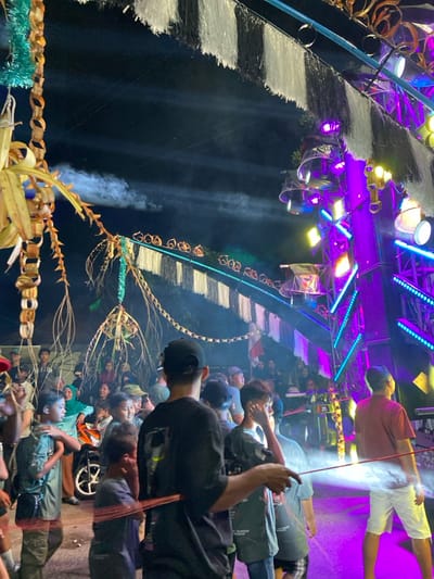 This image captures a nighttime outdoor event in Kedungbanteng, Indonesia. A crowd of individuals, predominantly males ranging from children to adults, is gathered, largely facing towards the right side of the frame. In the immediate foreground, a man wearing a black cap and t-shirt holds a red rope horizontally across the lower-middle of the image.

The scene is extensively illuminated by artificial lighting. On the right, a large vertical truss structure supports multiple stage lights emitting intense purple, blue, yellow, and white beams, alongside integrated speaker cabinets and several clear plastic-domed lighting fixtures. Above the crowd, a horizontal structure adorned with black and white fringed tassels and smaller, circular decorations spans the frame. On the left, elaborate woven palm leaf decorations, resembling chains and intricate abstract forms, hang alongside green tinsel.

Artificial smoke or fog is visible, catching and reflecting the stage lights. An Indonesian flag, red and white, is partially visible vertically in the background, center-right. A motorcycle is partially visible behind attendees on the left. Among the crowd, a person in a wheelchair is present on the left side.