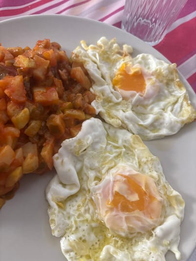A high-angle, close-up shot captures a white ceramic plate holding a prepared meal, situated on a red and white striped tablecloth, indicative of a domestic setting within Urbanización Encinares De Alcolea, Spain. The plate features two sunny-side-up fried eggs positioned on the right, each displaying a vibrant orange, liquid yolk surrounded by firm, opaque white albumen with slightly crisp, browned edges. To the left, a heap of diced, reddish-orange and greenish-yellow ingredients, primarily cubed potatoes and possibly bell peppers and tomatoes, are coated in a dense, reddish-brown sauce. A clear, fluted glass containing a transparent liquid is partially visible in the upper right background, resting on the striped surface.