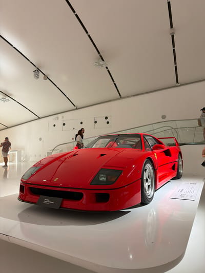 The image shows a red Ferrari F40 on display in a museum. The car is positioned in the foreground, angled slightly to the left, with its front end facing the viewer. The car's iconic design, including its aggressive front fascia, distinctive side vents, and prominent rear wing, is clearly visible. A plaque in front of the car reads "MUSEO ENZO FERRARI MODENA".
The background is a modern, minimalist museum interior with white walls and a white floor. The ceiling features a track lighting system with multiple spotlights directed at the display area. In the middle ground, to the left of the car, a man in a floral shirt and shorts is standing, seemingly taking a photo with his phone. Further back, a couple is visible near a railing, looking towards the car or other exhibits. To the right of the car, a woman is standing on a raised platform, partially obscured by the car. The overall impression is that of a well-lit, contemporary exhibition space dedicated to automotive history. The presence of the "Museo Enzo Ferrari Modena" plaque confirms the location as the Enzo Ferrari Museum in Modena, Italy.
