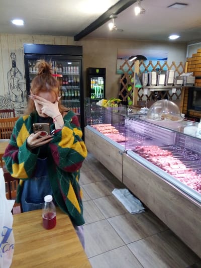 In Gol'yovo, Russia, a young woman with reddish hair, styled in a messy bun, is seen holding a smartphone in her left hand and covering her eyes with her right hand. She is wearing a green, yellow, and burgundy argyle-patterned cardigan over a dark top and denim overalls. A small bottle of red liquid sits on a wooden table in front of her. To her right, a refrigerated display case showcases raw meat skewers and cuts. Behind her, a refrigerator stocked with beverages stands against a wall adorned with a drawing of a wine bottle and grapes. The environment appears to be a food establishment, possibly a butcher shop or deli, with tiled flooring and a brick structure visible in the background.
