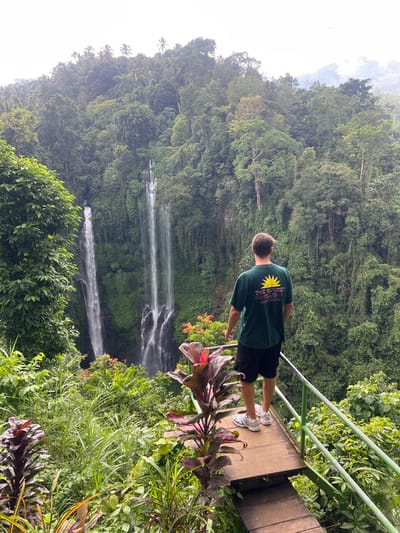 A person stands on a small viewing platform surrounded by lush greenery, gazing towards a multi-tiered waterfall cascading down a steep, tree-covered hillside. The person wears a dark green t-shirt, black shorts, and white sneakers. Metal railings provide a barrier along the platform's edge. Various plants frame the scene, adding to the tropical atmosphere. The sky is overcast.