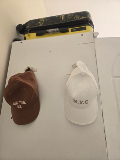 A yellow suitcase with black accents is resting on top of a white surface, possibly a cabinet or shelf. Two baseball caps are hanging from hooks on the vertical white surface below the suitcase. The cap on the left is brown and embroidered with "NEW YORK N.Y." in white lettering. The cap on the right is white and embroidered with "N.Y.C" in brown lettering. The environment appears to be indoors, with a plain wall in the background. There is no indication of the city of Deira, United Arab Emirates in the image.
