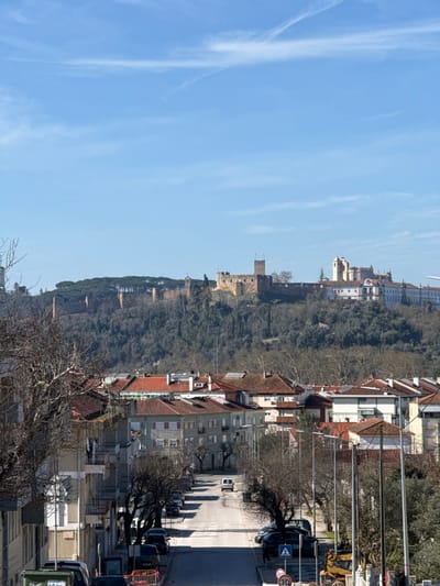 An outdoor, daytime image depicts a view in Tomar, Portugal, looking uphill from a residential street towards the Castle of Tomar and the Convent of Christ on a prominent forested hill.

In the foreground, a paved asphalt street ascends the incline, flanked on both sides by parked passenger vehicles. A white panel van is observed moving uphill in the center of the street. Multi-story residential buildings with terracotta-tiled roofs and balconies line both sides of the street. Bare deciduous trees and streetlights are spaced along the curbs. A green waste bin is visible in the lower-left corner. Traffic signage includes a blue pedestrian crossing sign and a circular "30" speed limit sign.

The mid-ground features a densely wooded hillside, primarily composed of bare deciduous trees with some interspersed evergreens, acting as a natural buffer between the residential area and the summit structures.

In the background, atop the hill, the historical complex of the Castle of Tomar and the Convent of Christ is prominently displayed. The Castle of Tomar exhibits light brown stone walls, battlements, and multiple towers, with flags flying from at least one tower. Adjacent to the castle, the Convent of Christ presents a series of lighter-colored, multi-story buildings with distinct architectural features. A dense line of dark green evergreen trees crowns the summit behind the castle and convent structures.

The sky above is clear blue with sparse, high-altitude white wispy clouds, including one distinct linear contrail-like cloud formation. No discernible individuals are engaged in specific actions within the visible frame.
