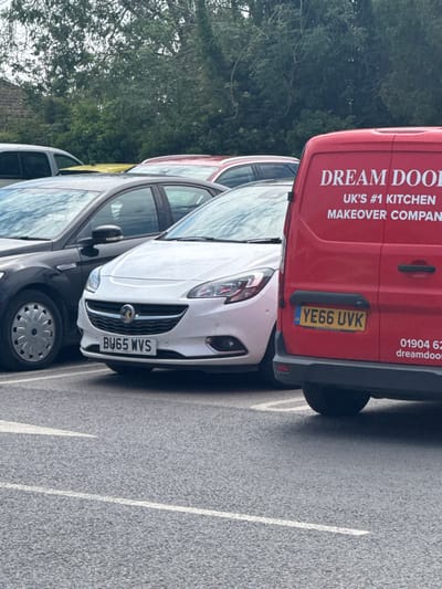 A red van branded with "DREAM DOOL UK's #1 KITCHEN MAKEOVER COMPAN" partially obscures a row of parked cars. The van's license plate reads "YE66 UVK." A white car is parked beside it, bearing the license plate "BU65 WVS." The van also shows the phone number "01904 62" and the website "dreamdoom." The vehicles are parked on asphalt within a parking area with white lines delineating the spaces, suggesting an outdoor setting with trees in the background.