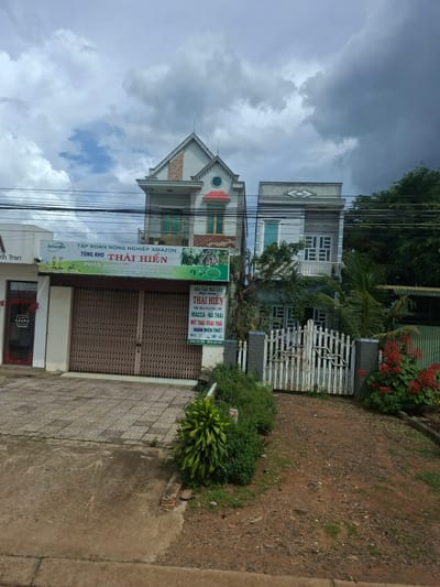 The image depicts a street scene in Tan Phu 2, Vietnam, with a focus on two adjacent buildings. The building on the left is a two-story structure with a closed metal gate at ground level, suggesting a shop or business. Above the gate, a large sign displays "Amazon TONG KHO THAI HIEN" and information about agricultural products. To the left of this building, a smaller section with a sign reading "AZURA" is visible. The building on the right is a taller, three-story structure with a white picket fence at ground level, indicating a residential or mixed-use property. The sky is overcast with dark clouds. The surrounding environment appears to be a moderately urbanized area, with trees and vegetation present. The ground is a mix of paved areas and dirt.