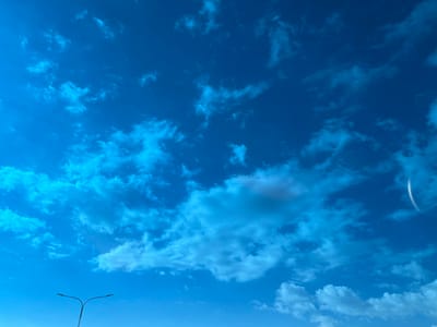 The image shows a clear blue sky with scattered white and light gray clouds. In the lower left corner, there is a street light. A bright white curved shape is visible in the upper right corner, potentially a lens flare.
There is no location-specific information available.
There are no people, subjects, objects, actions or interactions visible.
The environment is an open, clear sky.
