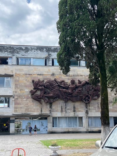 An exterior shot captures a large, multi-story building with a textured, stone-like facade in Pitsunda, Abkhazia. A prominent, dark, bas-relief sculpture depicting figures in a chariot, horses, and masks adorns the upper section of the building's wall. The building has numerous windows, some with white curtains, and appears to be in a state of disrepair in some areas, with peeling paint on the upper levels. To the right of the building, a tall, green tree with a moss-covered trunk stands prominently, partially obscuring the building. In the foreground, a paved area with grass and a planter box is visible. Two young people, one wearing a backpack, are walking on the paved area in front of the building. A portion of a silver car is visible in the lower right corner of the frame. The sky is overcast with visible clouds.