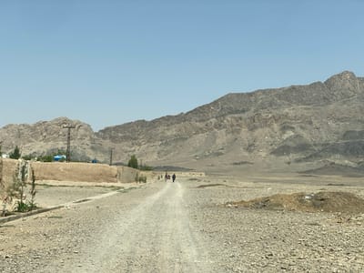 The image shows a dirt road in the desert of Panjwai, Afghanistan. The road leads up to a group of people walking in the distance. The road is surrounded by a rocky desert landscape with a large mountain range in the background. There are a few trees and shrubs growing in the area. There are power lines and poles along the side of the road. The scene is taken during the daytime under clear, sunny skies.  The coordinates provided place this location in a remote, arid area of Panjwai, Afghanistan, likely near a rural settlement.