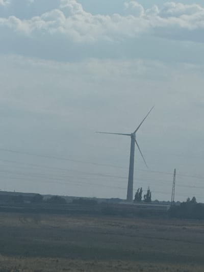An outdoor, slightly overcast scene features a wind turbine prominently in the center, with its blades angled. In the distance, a line of trees and industrial structures is visible, with a thin, tall tower to the right of the wind turbine. The foreground consists of a grassy, brownish field. The overall impression is one of a rural or semi-industrial landscape under a cloudy sky.