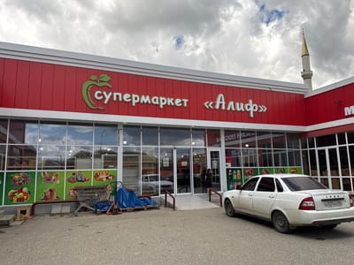 The image shows the exterior of a supermarket in the city of Mayrtup, Russia. The supermarket has a red facade with white trim and large glass windows. The name of the supermarket, "Супермаркет «Алиф»", is displayed in large green and white letters on the front. A golden minaret of a mosque is visible in the background.
In front of the supermarket, there is a white Lada car parked. There are also several shopping carts, a pallet with a blue tarp, and a small wooden structure with a black cat inside. Inside the supermarket, through the windows, one can see shelves stocked with goods and several people browsing. The sky is cloudy. The ground in front of the supermarket is paved with asphalt.