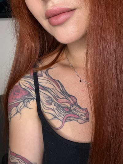 The image shows a close-up of a person with auburn hair, wearing a black tank top and a necklace. A tattoo of a dragon is visible on their shoulder and arm. The person's lips are painted with pink lipstick. The background is out of focus and appears to be an indoor setting. There is no discernible location in Oldham, United Kingdom.