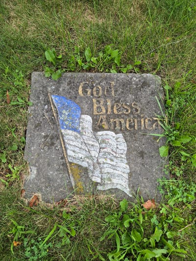 The image shows a flat, weathered stone embedded in the ground, surrounded by green grass and some scattered dry leaves. The stone features a painted design of the American flag with the words "God Bless America" inscribed above it. The flag's colors are faded, with white and blue sections for the stripes and stars, and red accents. The lettering is in a bold, gold-colored font. The scene appears to be outdoors, likely in a park, cemetery, or a residential yard, given the natural setting and the presence of the stone marker. The lighting suggests it is daytime, with no specific cues about the weather, though the lush grass might indicate a season with sufficient rainfall. The overall mood is patriotic and contemplative. The text "God Bless America" is clearly visible on the stone.