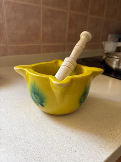 The image shows a yellow ceramic mortar with a wooden pestle inside. The mortar has a flared rim and some green discoloration on its surface. The pestle is cylindrical with a rounded end. The mortar and pestle are sitting on a light-colored countertop, possibly in a kitchen. The background includes brown tile flooring and a glimpse of a stainless steel pot or pan. While it's difficult to definitively confirm, the location is likely a residential setting within Urbanización Encinares De Alcolea, Spain, based on the provided information.