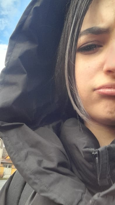 An outdoor portrait photograph captures the right side of a young female subject, positioned at a high angle. The setting is stated to be Meistratzheim, France. The subject is wearing a dark-colored, hooded jacket, with the hood pulled up and covering the upper left portion of the image. Her dark hair, featuring prominent silver-grey streaks, frames her visible cheek. Her right eye is partially visible, beneath a thick, dark eyebrow. Her lips are closed and downturned, forming a pouting expression. A zipper detail is visible on the right front of the jacket. The background is blurred, revealing residential buildings: a light-colored structure with a visible window opening, and a yellow-façade building with white window frames and bare tree branches nearby. Above the jacket's hood, a segment of blue sky with scattered white clouds is visible.