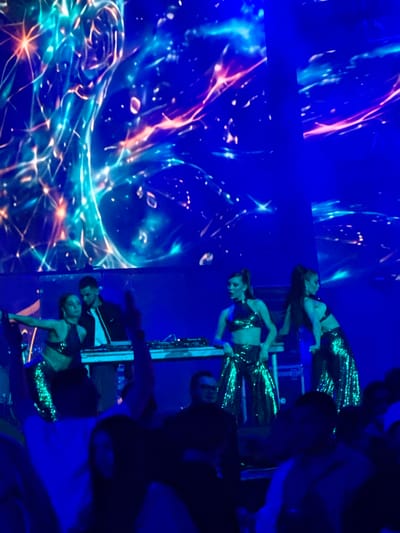 This image depicts a nighttime event in Tsilkani, Georgia. In the foreground, a crowd of people are visible, many of whom are out of focus. In the mid-ground, three women in sequined outfits are performing on a stage. Two of the women are wearing halter tops and shimmering pants, while the third woman is wearing a similar top and what appears to be a skirt or more pants. Behind the performers, a DJ booth is set up with a DJ. The background features a large, dynamic digital screen displaying a vibrant, abstract blue and orange light show. The overall atmosphere appears to be that of a concert or club performance.