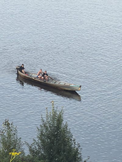In the city of Cherdyn, Russia, a long, narrow wooden boat with a small outboard motor is on the water. Four individuals are in the boat. One person, wearing a black shirt and a dark cap, is seated at the stern, appearing to operate the motor. Three other individuals are seated in the middle and forward sections of the boat. One person in the middle is wearing blue shorts and appears to be looking towards the right. The other two individuals are wearing dark shirts and are seated closer together. The water surface is rippled, suggesting a gentle breeze. In the foreground, a portion of a tree with green foliage and a few yellow wildflowers is visible. The overall scene depicts a leisurely activity on the water.
