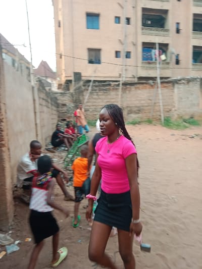 A woman in a pink shirt and black skirt walks on sandy ground. In the background, people are gathered near a wall. A child in an orange shirt and another in a white top and black skirt are also present. Tall buildings are visible behind the wall. The scene appears to be in a densely populated area.