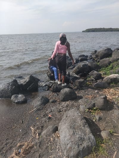 The image depicts a rocky shoreline scene in West Uyoma ward, Kenya, under a bright, overcast sky. A large body of water, possibly a lake, extends to the horizon, showing slight ripples. In the foreground, two individuals are positioned among dark, coarse rocks at the water's edge.
One individual, seen from behind, is standing upright facing the water. This person has dark skin, wears a pink long-sleeved crop top, dark trousers, and white athletic shoes. A patterned headwrap covers their hair, which appears dark and braided. Their left arm is slightly extended over the water, with the hand open.
Adjacent to this individual, to their left, a second person is partially visible, obscured by rocks and the first individual. This person is wearing a dark top and blue trousers, and appears to be crouching or bending forward, interacting with the water or submerged rocks.
The immediate shoreline consists of dark grey to black angular rocks of varying sizes, interspersed with dark, sandy/muddy ground. Sparse green vegetation grows among the rocks and on the ground. Visible debris on the dark ground includes dried fibrous material and small fragments of refuse. In the distance, across the expanse of water to the right, a tree-covered landmass, consistent with an island or distant shore, is visible.
