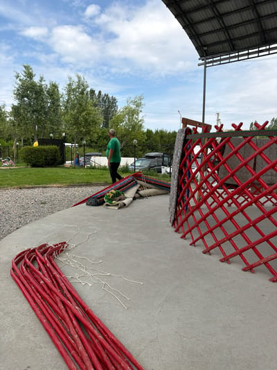 In the city of Kyzyl-Tuu, Kyrgyzstan, a man in a green shirt and a patterned cap walks away from the camera on a concrete surface. In the foreground, a bundle of red, curved poles with white strings is laid out on the concrete. To the right, a portion of a red, latticed structure, possibly part of a yurt frame, is visible. Stacked on the concrete behind the poles are several rolled-up rugs of various colors and patterns. In the background, there is a grassy area with trees and a black fence. Beyond the fence, several vehicles are parked, including a black SUV. The sky above is blue with scattered white clouds.
