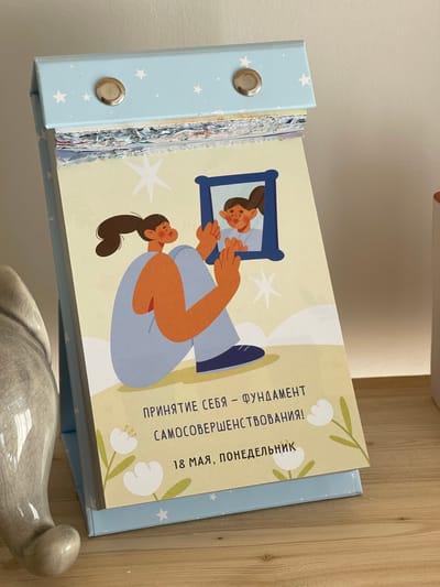 On a wooden surface, a light blue calendar with white stars is shown. The calendar is open to a page with a drawing of a person looking in a mirror. The text on the page reads "Принятие себя – фундамент самосовершенствования! 18 мая, понедельник" which translates to "Acceptance of self - the foundation of self-improvement! May 18, Monday." The bottom of the calendar sits on what appears to be the back of a ceramic gray cat. The location is unknown.
