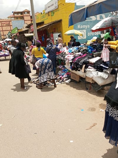 The image depicts an active outdoor street market located in Tala ward, Kenya. A paved road occupies the foreground and mid-ground, leading to a bustling commercial area.

Along the right side of the road, multiple market stalls are set up, operating under blue tarps and a small yellow-and-white umbrella for shade. These stalls display large quantities of merchandise, primarily piles of clothing and assorted footwear, including sandals, arranged on tables and directly on the ground. Specific objects visible include a cardboard box labeled "UMOJA" and a black shopping bag with the brand "LULU".

In the mid-ground, several individuals are engaged in market activities. Three women are prominent: one in a black coat stands facing the stalls, another in a yellow top is observing items, and a third woman in a blue and white patterned dress is bent over, examining shoes placed on the ground. Another individual wearing a blue shirt with an "Africom" logo stands near the merchandise. On the far right, the lower body of a person wearing a dark blue pleated skirt is partially visible. Further back, numerous other people are present, indicating a busy shopping environment.

The background consists of a row of commercial buildings. A prominent yellow building displays a "SWISA STORES" sign. An adjacent light blue building features signage partially visible, including the word "Kenya" and a phone number starting "0711 066". A utility pole stands centrally in the mid-background. The scene is illuminated by natural daylight.