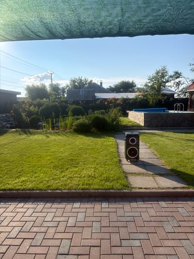 The image depicts a backyard scene in Chernousovo, Russia, on a sunny day. In the foreground, a paved walkway made of reddish-brown and grey interlocking bricks leads towards a lush green lawn. A tall, black portable speaker with blue accents stands on a stone path dissecting the lawn.

The middle ground features a well-maintained garden with various shrubs, including rounded evergreen bushes, and tall green reeds growing around a small pond. Orange flowers bloom in the garden, adding a splash of color. Beyond the garden, a stone-walled swimming pool with a blue cover and a ladder is visible.

In the background, several residential buildings with grey, corrugated roofs are scattered amidst trees. Power lines and a utility pole are also present. The sky is a clear, bright blue with a few wispy white clouds. A green mesh shade cloth is stretched across the top of the image, partially obscuring the sky. The overall impression is that of a serene, well-kept outdoor living space.