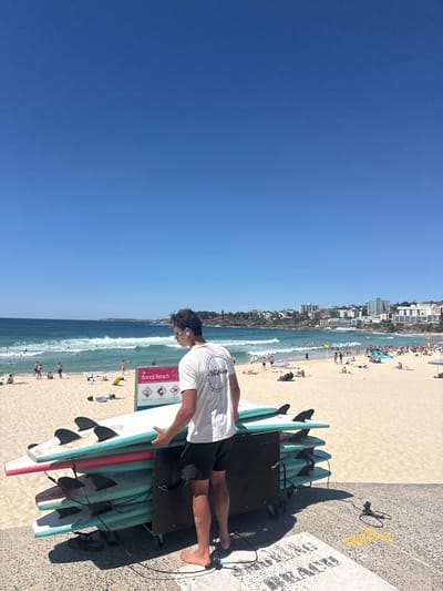 A man stands next to surfboards, wearing a white t-shirt and dark shorts. The surfboards are light blue and pink. The man is on a concrete surface next to a sandy beach, with buildings along the shoreline. The sky is bright blue. People are on the beach and in the water. A sign is visible on the left side of the frame, near the surfboards.