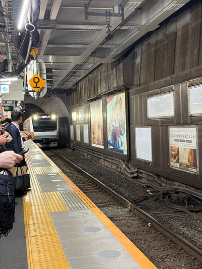 The image depicts a subway station platform in Bunkyō, Japan, with a train approaching on the tracks. On the platform, several individuals are waiting, one of whom is holding a mobile phone. The platform is marked with yellow tactile paving to guide visually impaired passengers. Adjacent to the tracks, a series of advertisements are displayed on the station wall. The station ceiling shows exposed infrastructure, including pipes and lighting fixtures. A yellow diamond-shaped sign with a black symbol is visible on a pole, a common visual indicator in Japanese train stations. The overall environment is that of a busy urban transit hub.