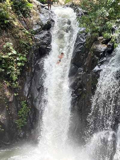 A natural waterfall scene in Ambengan, Indonesia, depicts a recreational activity. A young male, clad in dark swimming shorts, is observed sliding head-first on his back down a central channel of turbulent white water. His arms are bent, and his head is partially submerged but visible above the water. The lower legs and feet of a second individual are visible at the top edge of the waterfall, standing on dark rock. The waterfall is contained within steep, dark rock formations covered by dense green foliage, including trees and vines. Water cascades forcefully, creating significant spray and foam at its base.