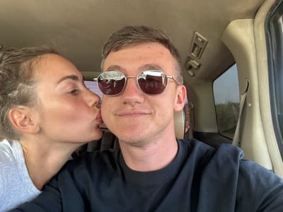 The scene depicts two individuals inside a vehicle, located in Paje, Tanzania. A female subject, positioned on the left, has light brown hair pulled back and wears a grey short-sleeved top. She is captured mid-action, kissing the right cheek of the male subject. The male, positioned centrally, wears silver-rimmed round sunglasses with reflective lenses and a dark long-sleeved shirt, displaying a subtle smile towards the viewer. The vehicle's interior features a light tan upholstered ceiling, an overhead air vent, and a grab handle. Through the vehicle's right-side window, blurred green vegetation is visible, indicating an outdoor environment. The interaction between the subjects is an affectionate gesture within the confines of the vehicle.