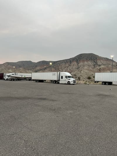 The image shows a row of semi-trucks parked on an asphalt lot. The trucks are white and have long trailers. One truck in the foreground has the text "Premier" visible on its trailer. In the background, a mountainous landscape is visible with striated rock formations. There are also streetlights present in the scene. The sky is overcast and appears to be either early morning or late evening. The overall scene suggests a truck stop or rest area. No people are visible in the image. The location is identified as Salina, United States of America.