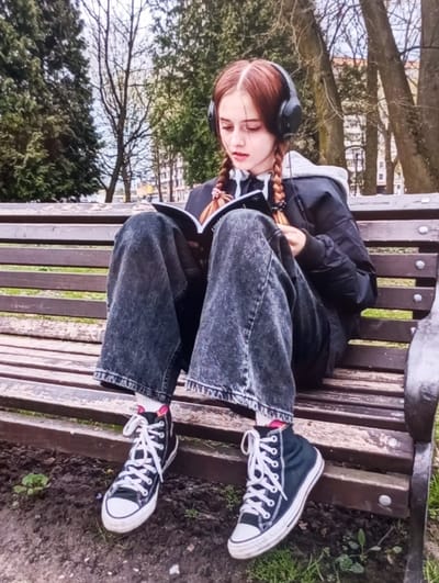 A young female with reddish-brown hair, styled in two braids secured with black elastic ties, is seated centrally on a brown wooden park bench in an outdoor setting in Keskanterak, Uzbekistan. She wears black over-ear headphones and is actively reading an open black notebook or book held in her lap. Her attire consists of a black bomber-style jacket worn over a light grey hooded sweatshirt, loose-fitting dark wash jeans, white ankle socks, and black high-top canvas sneakers with white laces, white rubber toe caps, and white soles. The background features a park environment with numerous trees, some deciduous with bare branches and some evergreen, under an overcast sky. The ground immediately beneath the bench is visible as dark soil with sparse green vegetation.