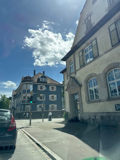 The image depicts a street scene in Nürtingen, Germany, on a sunny day with scattered clouds. In the foreground, the rear of a black car is visible on the left side of the frame. The street curves to the right, and a sidewalk runs alongside it.

To the left of the street, a dark blue building with white-trimmed windows and white shutters is prominent. This building has a distinctive octagonal section on its left side. A traffic light on a pole shows a green signal. Several people are standing on the sidewalk in front of this building.

To the right of the street, a light-colored building with arched windows and a decorative facade is visible. This building has shutters on its windows and appears to be an older structure. A sign with the word "GO!" is partially visible above an entrance.

The sky is blue with fluffy white clouds. The overall impression is of a calm urban environment on a clear day.