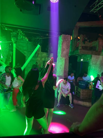 The image depicts a nighttime scene inside a dimly lit establishment, likely a nightclub, in Canet-en-Roussillon, France. Green and purple spotlights illuminate the area, casting vibrant hues on the patrons and the decor. In the foreground, a woman with long dark hair is captured from behind, wearing a black top and black shorts, her left arm raised. To her left, a man in sunglasses and a plaid shirt over a white tank top is smiling, with a woman in a white top and red pants standing beside him. Other individuals are visible throughout the scene, some seated, others standing and socializing. The environment features textured walls, possibly designed to resemble ruins or a jungle theme, with foliage and exposed structural elements. A large blue cooler labeled "GREY GOOSE" is placed on a wooden crate in the background. The atmosphere suggests a lively social gathering with music and dancing.