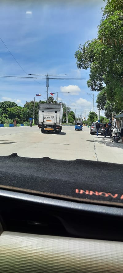The image shows a street scene in Imus, Philippines, under a bright, partly cloudy sky. In the foreground, the textured interior of a vehicle is visible, including a dark mat with the word "INNOV" partially visible in red lettering. The road ahead is paved and appears to be a multi-lane intersection or a wide road with traffic.

Dominating the mid-ground is a white box truck, driving away from the camera. The truck has Philippine flags mounted on its roof, fluttering in the breeze. To the left of the truck, a lone motorcyclist is also traveling on the road. Further down the road, a blue tricycle is visible, as well as a gray sedan with people standing near it and motorcycles parked on the side.

The environment is characterized by lush green trees lining the sides of the road and utility poles with numerous wires stretching across the scene. The overall impression is one of a typical day in a busy urban or suburban area of Imus.