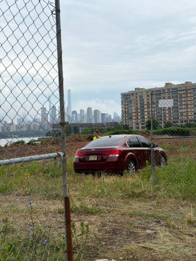 In North Bergen, New Jersey, USA, a red Subaru Legacy sedan is parked in an overgrown lot. The car is facing right, with its rear end visible. A person wearing a yellow safety vest stands near the car. In the background, a cityscape with numerous tall buildings and a river is visible under a cloudy sky. A chain-link fence with rusted poles runs through the foreground of the image, partially obscuring the scene. The ground is covered with dry grass and some small blue wildflowers.