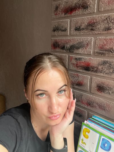 In this close-up, self-taken photograph, a young woman with light brown hair pulled back from her face is positioned in the foreground, resting her chin on her right hand. Her light blue eyes are looking directly at the camera, and her lips are slightly pursed. She is wearing a black t-shirt, a silver necklace, and a black smartwatch on her left wrist. A small nose piercing is visible. To her right, a stack of colorful children's alphabet books is partially visible, with pages showing illustrations of a caterpillar and the letter "C" for "cat" and "cake," and the letter "D" for "donut." The background consists of a textured, light brown wall on the left and a decorative brick wall with a distressed finish in shades of white, gray, black, and red on the right. The lighting appears to be natural, casting soft shadows. The setting is an indoor environment, likely a room in a residential building in Zarichne, Ukraine, given the context.