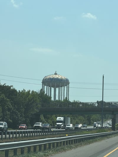 The image shows a water tower with the United States Air Force logo and the word "FORCE" visible on its side. The water tower is a prominent structure in the background, set against a clear blue sky with a few wispy clouds. It is supported by a series of tall, thin legs. In the middle ground, a highway overpass with traffic is visible, with trees and vegetation lining the sides of the road. Several cars and trucks are driving on the highway. The foreground features a guardrail and some dry grass. The scene appears to be a roadside view in Forestville, United States of America.