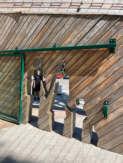 A person stands on a wooden structure that appears to be a tiered seating area or a playground. The structure is made of diagonal wooden planks with some green metal supports and brackets visible. The person is wearing a black t-shirt and black shorts and is looking at the camera. The area is illuminated by sunlight, casting shadows on the wooden planks and the gray tiled ground next to the structure. In the background, a red car and a white air conditioning unit are visible. A green metal fence with a grid pattern is in the foreground. The overall scene suggests an outdoor setting, possibly a park or a recreational area. The time of day appears to be daytime, and the weather seems to be sunny. The person's expression is neutral. There is some faint text on the person's t-shirt, which is partially obscured.