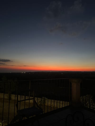 The image captures a twilight scene from a balcony overlooking the city of Ragusa, Italy. The sky is a gradient of deep blue at the top, transitioning through lighter blues and purples to a vibrant band of orange and red along the horizon, indicating the sunset or sunrise. A single bright star is visible in the upper left portion of the sky. Below the horizon, the landscape is dark, with scattered lights suggesting the urban area of Ragusa. In the foreground, a black metal railing runs horizontally across the balcony. To the left, a wooden chair is positioned behind the railing. To the right, a stone pillar or balustrade is visible. The balcony floor is paved. The overall atmosphere is serene and twilight.
