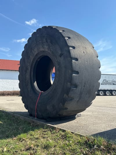 A large, heavily worn, dark gray industrial off-the-road (OTR) tire is standing upright on a paved asphalt surface. The tire rests on two dark, rectangular blocks, and a red strap, possibly a securing strap, hangs from its lower interior rim towards the ground. To the immediate left, a strip of green grass with sparse brown patches is separated from the pavement by a concrete curb. In the background to the left, a white building with a red tiled roof is visible. To the right, a white semi-trailer truck displaying "Bussing Transporte" is parked. The scene is outdoors under a clear blue sky with scattered white clouds. This installation is located in Weißenburg in Bayern, Germany.