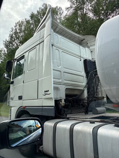 The image is a photograph of a white DAF truck with a large cylindrical tank trailer, viewed from the side of another vehicle. The truck cab is prominent, showing the driver's side door, mirrors, and the DAF logo. The rear of the truck cab reveals mechanical components, including coiled air hoses and a black radiator-like unit. The tank trailer is a large, white, cylindrical structure. The background consists of a cloudy sky and lush green trees. In the foreground, a side mirror of the vehicle from which the photo is taken reflects the interior with a person inside, and the road with trees. The scene appears to be on a paved road, likely a highway, surrounded by nature. There is no indication of the city of Акцябрскі сельскі Савет, Belarus within the image content.