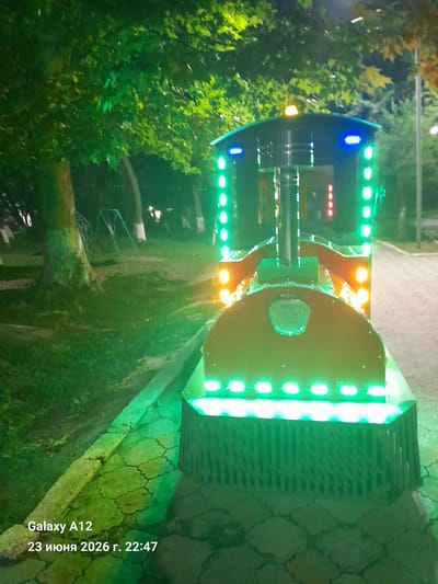 This is a photograph of a brightly lit, toy train attraction, likely in a park or amusement area, in Kara-Köl City, Kyrgyzstan. The train is adorned with numerous green and blue LED lights, with some orange lights also visible. The train appears to be made of metal and plastic, with a large, round section dominating the foreground and a smaller section extending to the left. The setting suggests an outdoor environment, with trees and foliage visible to the left of the train, and a paved walkway or path behind it. The ground appears to be covered in grass and a patterned stone surface.
The lighting indicates that the photograph was taken at night or during twilight. The presence of the train suggests an attraction that operates during these hours. The overall mood is festive and colorful due to the vibrant lights.
Visible text in the image reads: "Galaxy A12" and "23 июня 2026 г. 22:47", indicating the device used to capture the image and the date and time it was taken. There is no visible text on the train itself. No people are visible in the frame. The weather appears to be clear.