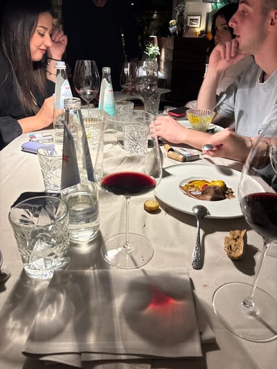 Captured indoors at what appears to be a restaurant, possibly in Karneid - Cornedo all'Isarco, Italy, the photograph presents a scene of dining. Two individuals are distinctly visible at the table: one pouring water from a glass bottle and another seen near a plate with food. A male diner seems to be eating or preparing to eat, with a fork and spoon visible near the plate. The table is set with numerous wine glasses, some filled with red wine, a water bottle, and textured drinking glasses. The food on the plate includes what appears to be dessert, with distinct colours suggesting different components like chocolate and fruit. The setting suggests a warm, possibly dimly-lit environment, typical of an intimate restaurant setting.